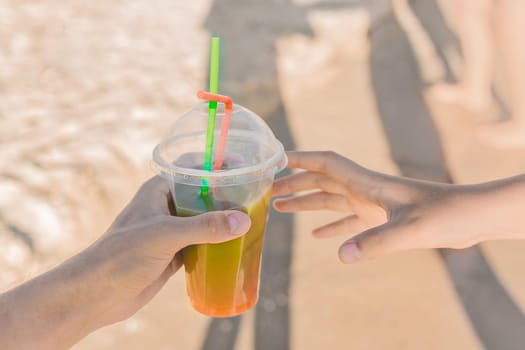 The guy's hand passes a colored chilled non-alcoholic cocktail into the girl's hand against the background of sea beach.