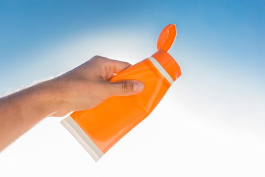 The guy's hand holds sunscreen against the background of a blue sky and a sea beach.