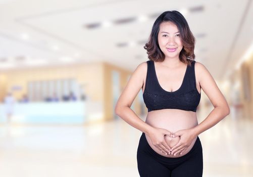 Pregnant Woman holding her hands in a heart shape on her belly in hospital background