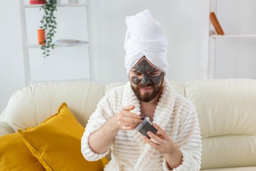 Bearded man having fun with a cosmetic mask on his face made from black clay. Men skin care and spa at home.