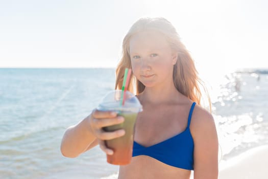 A young girl with blonde hair of European appearance, a teenager holds a colored cold non-alcoholic cocktail in her hand against the background of the sea beach.