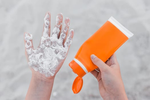 The girl holds sunscreen, in her hand and squeezes it on her arm palm to protect her from the sun against the background of beach sand.