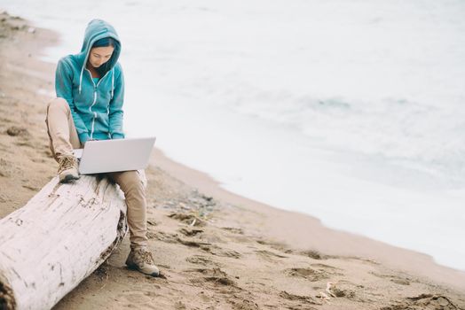 Freelancer girl sitting on tree trunk and working on laptop on beach near the sea. Freelance concept