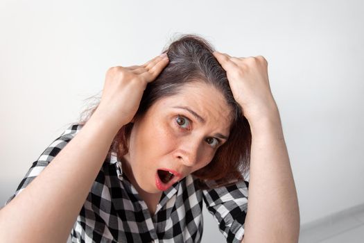 Horrified young woman looking in the bathroom mirror staring open mouthed at the first grey hair on her scalp, a first sign of ageing, or noticing that she is suffering from dandruff