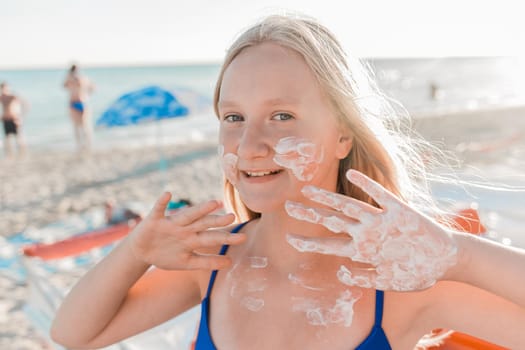 Portrait of a young positive teenage girl blonde of European appearance with sunscreen on her face and body against the background of a sea beach.