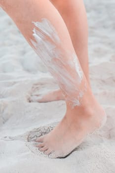 The legs of a young girl stand on the beach white sand with sunscreen on the skin.