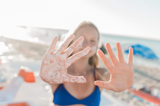 A young European teenage girl stretched her arms forward with sunscreen and sun protection on her palms against the backdrop of a sea beach.