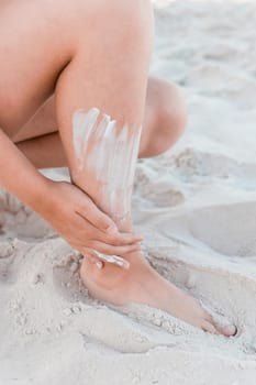 A young girl's hand smears sunscreen on her leg next to the beach sand.