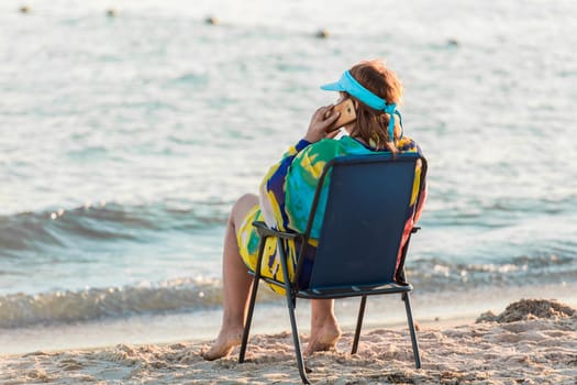 An adult woman in a pareo sits on a folding chair by the sea water on the shore and talks on a smartphone.