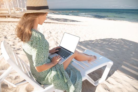 Rear view of an attractive young Caucasian woman in a green dress and hat sits on a lounger with a laptop in her hands in the shade of a wooden umbrella. Cut out laptop screen.