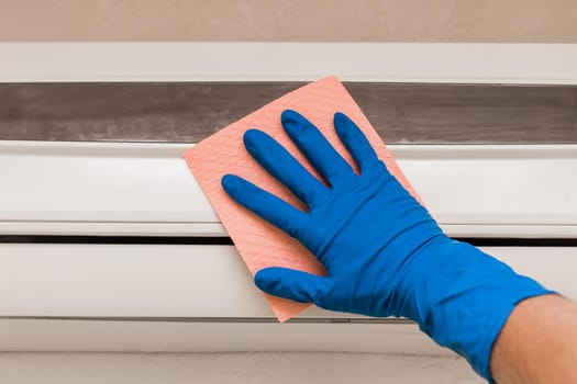 The hand of a man in a blue rubber household glove wipes and cleans the air conditioner. Maintenance and cleaning indoor service.