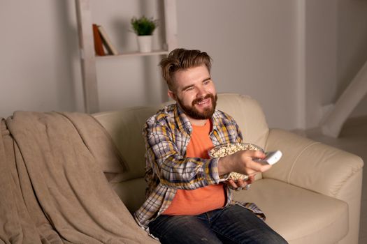 Portrait young man sitting on couch and watching TV holding remote and eats popcorn
