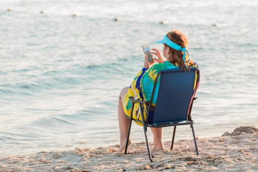An adult woman in a pareo sits on a folding chair by the sea water on the shore, and sitting in a smartphone or mobile phone.