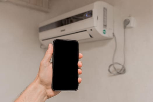 A person's hand holds a mobile phone or smartphone with a black screen for text and design, against the background of an air conditioner on the wall in a room of the house.