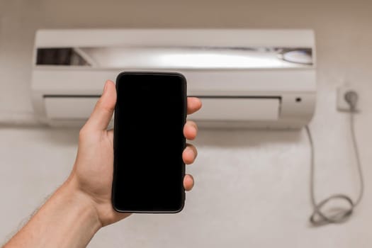 A person's hand holds a mobile phone or smartphone with a black screen for text and design, against the background of an air conditioner on the wall in a room of the house.