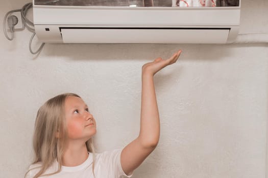 Young cute girl teenager blonde European appearance brings his hand to the air conditioner on the wall to check the heat and cold, the temperature in the room.