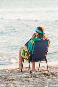 An adult woman in a pareo sits on a folding chair by the sea water on the shore, and talks on a smartphone or mobile phone.