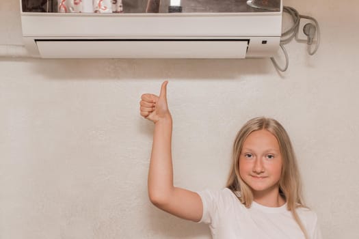 Young cute girl teenager blonde European appearance shows a cool thumb, looks into the camera and smiles next to the air conditioner on the wall in the room.