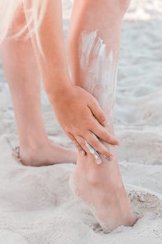 A young girl's hand smears sunscreen on her leg next to the beach sand.