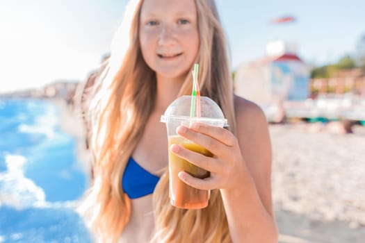 A young girl with blonde hair of European appearance, a teenager holds and drink a colored cold non-alcoholic cocktail in her hand against the background of the sea beach.