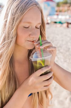 A young girl with blonde hair of European appearance, a teenager holds and drink a colored cold non-alcoholic cocktail in her hand against the background of the sea beach.