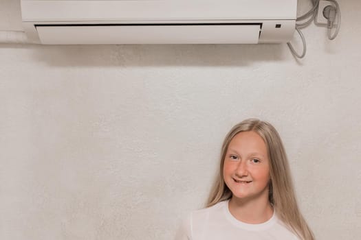 Cute young girl teenage blonde European appearance poses looking into the camera standing under the air conditioner on the wall in the room.