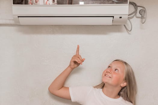 Cute teenage girl blonde European appearance points a hand finger at the air conditioner on the wall in the room.