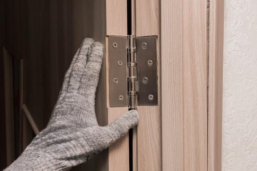 The hand of a working man in a construction glove touches and examines a wooden door opening with a door hinge, close-up. Installation or adjustment of metal parts.