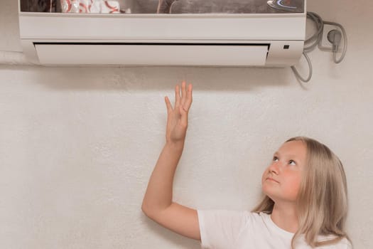 Young cute girl teenager blonde European appearance brings his hand to the air conditioner on the wall to check the heat and cold, the temperature in the room.