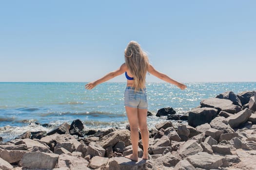 A young, teenage girl with long hair blonde stands on the rocks near the sea shore on the beach spreading and raising her hands to the sides.