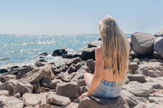 A young teenage girl with long blonde hair blonde sits on the rocks of the breakwater near the sea shore on the beach.