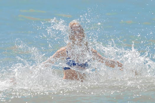 Young cheerful girl teenager blonde European appearance makes splashes of water with her hands in the sea.