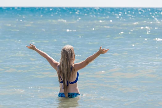 A young teenage girl with long blonde hair raised and spread her arms to the sides against the background of the sea water and horizon line. The concept of freedom.