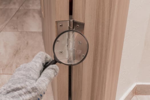 The hand of a male worker in a construction glove examines through a magnifying glass the door hinge of a wooden door. The concept of installation and fixation of metal parts.