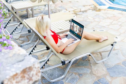 Woman Using Laptop Whilst Relaxing On Sun Lounger By Pool.