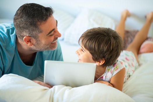 Funny dad and lovely little girl are having fun on the bed. Middle-age father with her eight years daughter using digital tablet in bedroom.