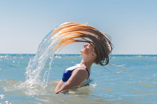 Young attractive girl teenager of European appearance throws wet, long hair up into the sea against the background of the horizon line and blue sky.