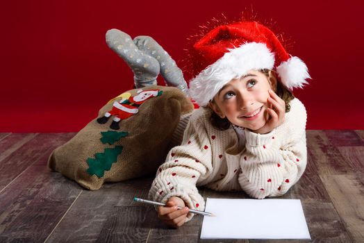 Adorable little girl wearing santa hat writing Santa letter on wooden floor. Winter clothes for Christmas.