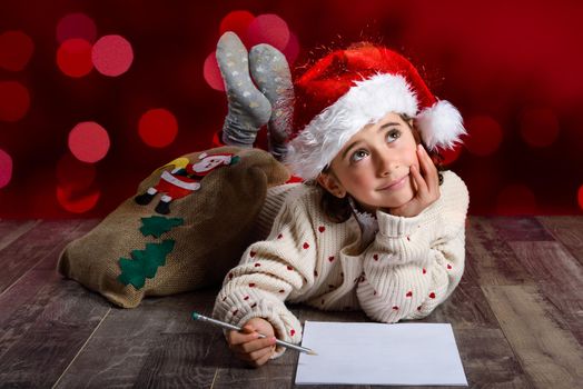 Adorable little girl wearing santa hat writing Santa letter on wooden floor. Winter clothes for Christmas. Red bokeh at the background