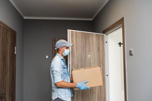 Delivery. Cheerful young courier holding a cardboard box while standing at the entrance of apartment.