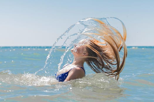 Young attractive girl teenager of European appearance throws wet, long hair up into the sea against the background of the horizon line and blue sky.