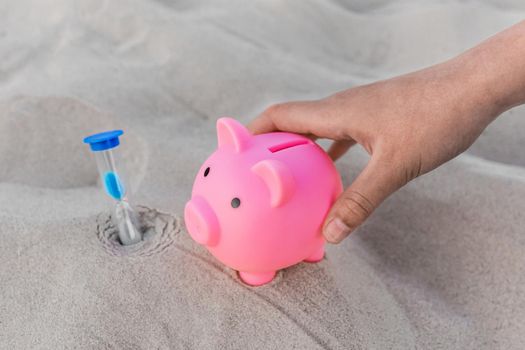 A human hand takes a piggy bank next to an hourglass against the background of white beach sand. Money, investments and fanance deposits for holidays at sea concept.