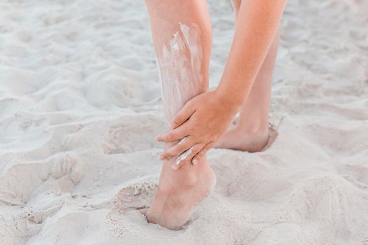 A young girl's hand smears sunscreen on her leg next to the beach sand.