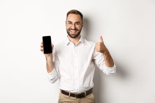 Satisfied business man in glasses showing thumbs up and demonstrating mobile phone screen, recommending app, standing over white background.