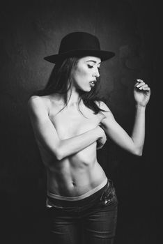 Topless woman wearing blue jeans, panties and hat on black background. Black and white studio photograph.