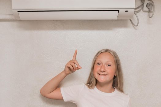 Cute teenage girl blonde European appearance points a hand finger at the air conditioner on the wall in the room.