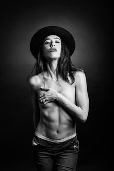 Topless woman wearing blue jeans, panties and hat on black background. Black and white studio photograph.