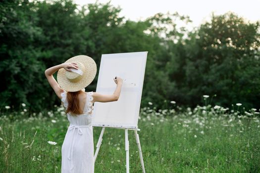 A woman in a white dress in a field with flowers paints a picture. High quality photo