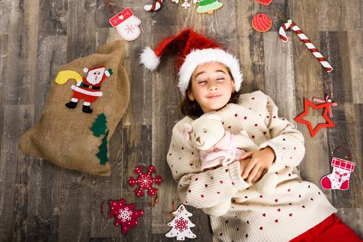 Adorable little girl wearing santa hat sleeping on wooden floor with Christmas ornaments. Winter clothes.