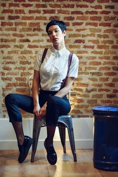 Beautiful young woman, model of fashion, with very short haircut, looking at camera sitting on chair, with a brick wall in the background. Beauty and hairstyles concept.